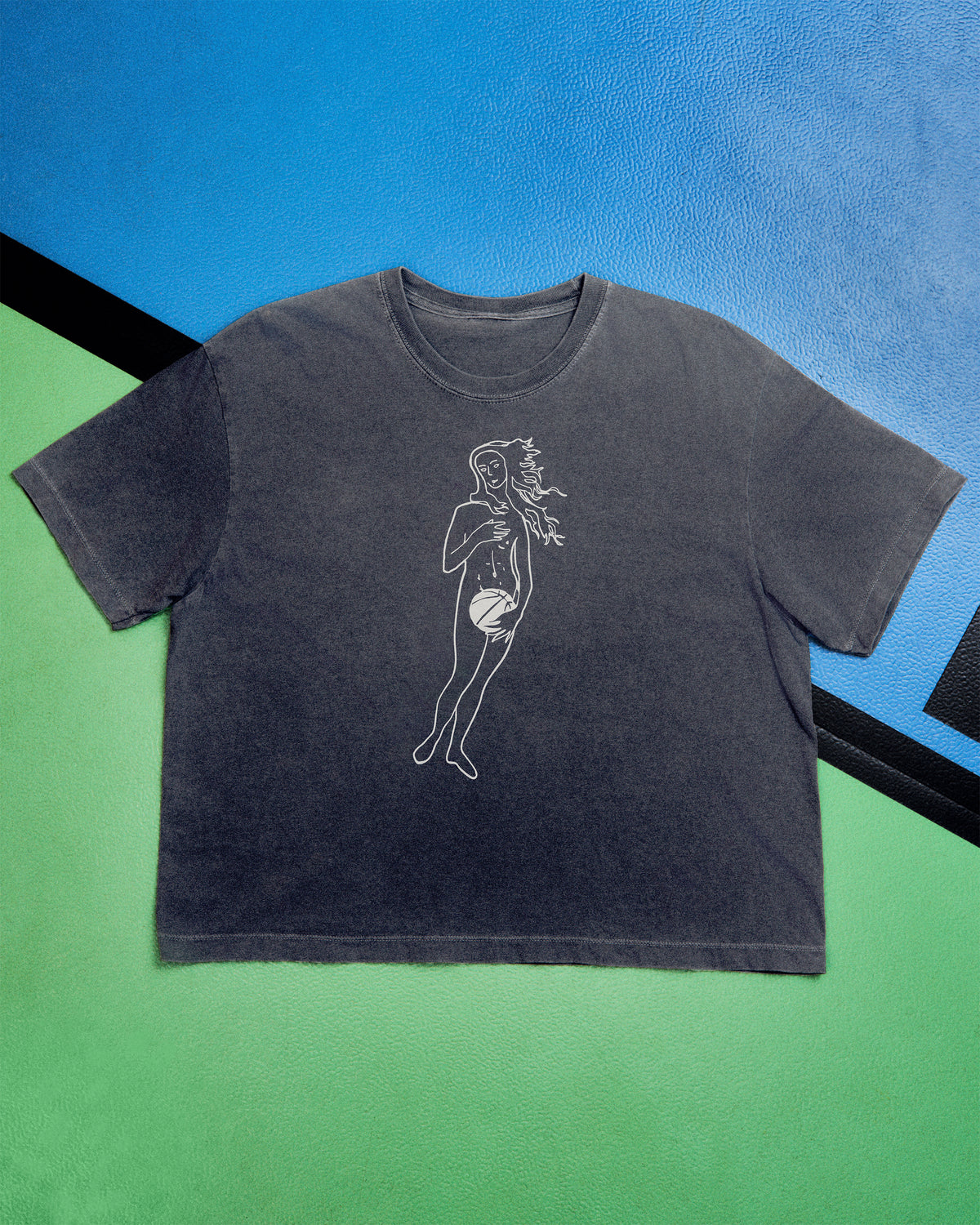 A dark gray washed short-sleeve t-shirt with a white line art graphic of a woman holding a basketball, laid flat on a split green and blue background.